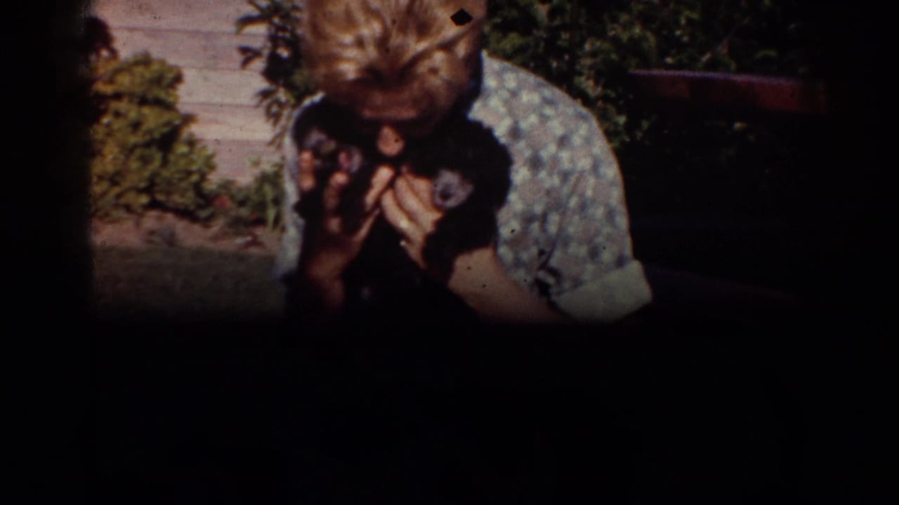 A 1962 Yorba Linda home-movie frame shows the woman holding the puppy close, with yard plants and outdoor furniture partly visible behind her.