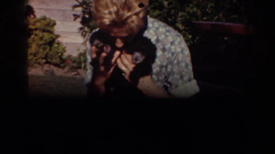 A 1962 Yorba Linda home-movie frame shows the woman holding the puppy close, with yard plants and outdoor furniture partly visible behind her.
