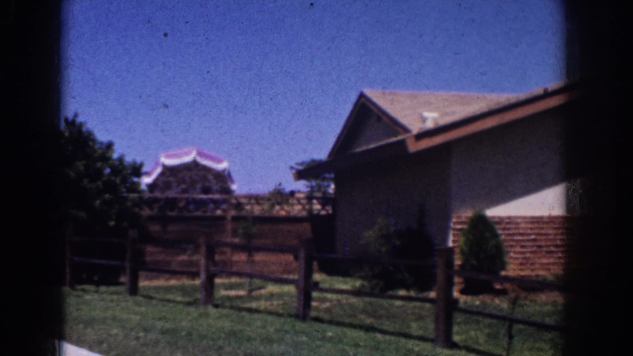 1962 Yorba Linda home-movie frame showing a ranch-style exterior, wood fence, lawn, shrubs, and a distant pagoda-like roofline.
