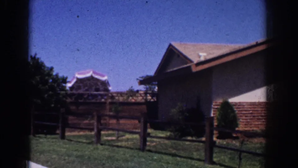1962 Yorba Linda home-movie frame showing a ranch-style exterior, wood fence, lawn, shrubs, and a distant pagoda-like roofline.