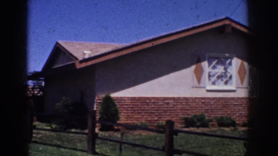 1962 Yorba Linda home-movie frame showing the ranch house side with brick foundation, decorative shutters, lawn, and fence.