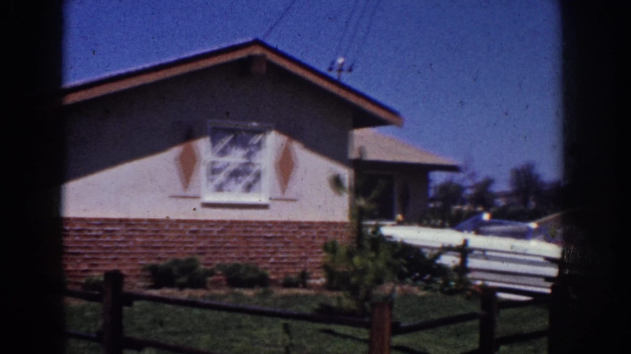 1962 Yorba Linda home-movie frame showing a cottage exterior, wooden fence, front lawn, and a pale car partly hidden in the driveway.