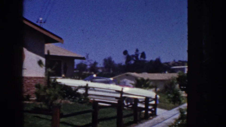1962 Yorba Linda frame emphasizing the driveway, pale parked car, wooden fence, lawn, sidewalk, and edge of the cottage.