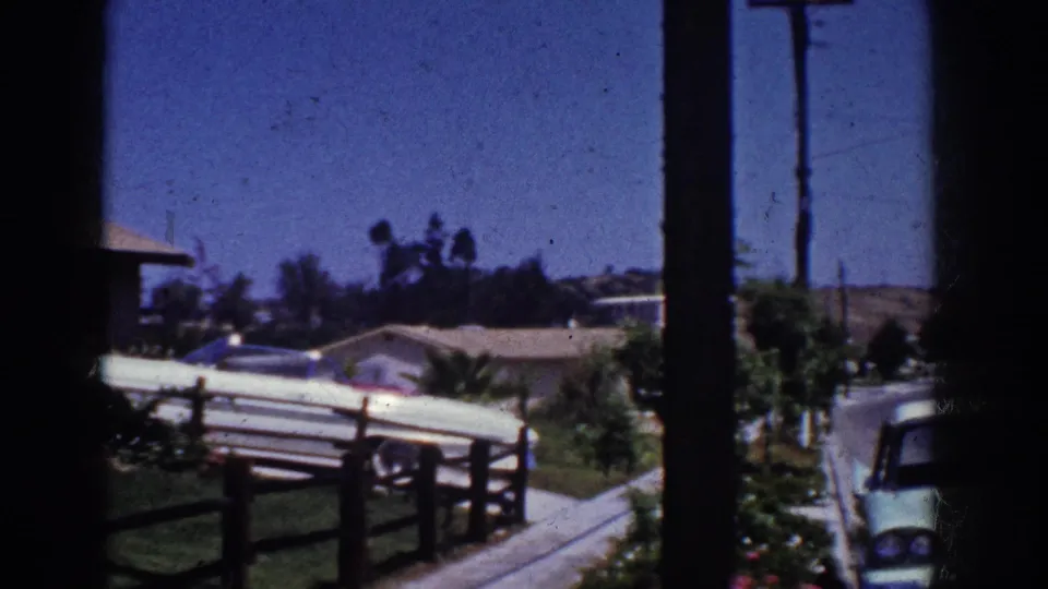 1962 Yorba Linda home-movie frame showing the driveway car, fence, sidewalk, utility pole, shrubs, and a partial vehicle at the edge.