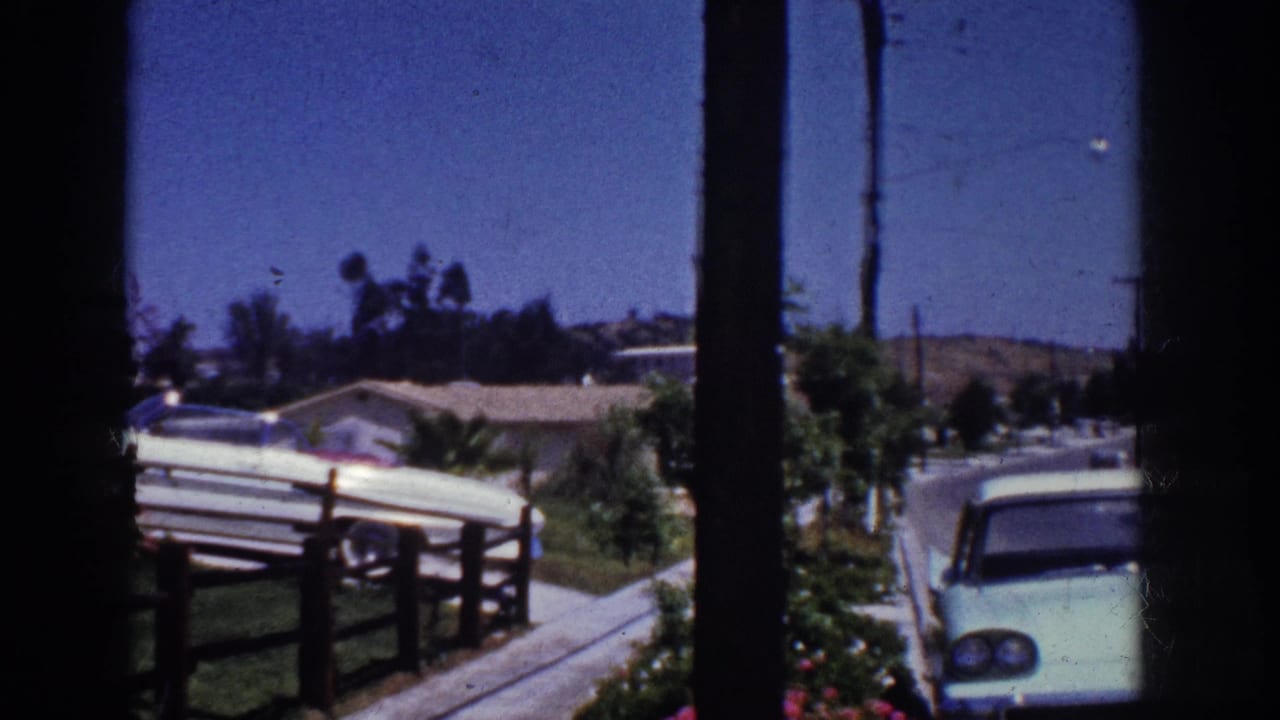 1962 Yorba Linda suburban street view through a window, showing a light blue sedan, low fencing, lawns, and houses under a blue sky.