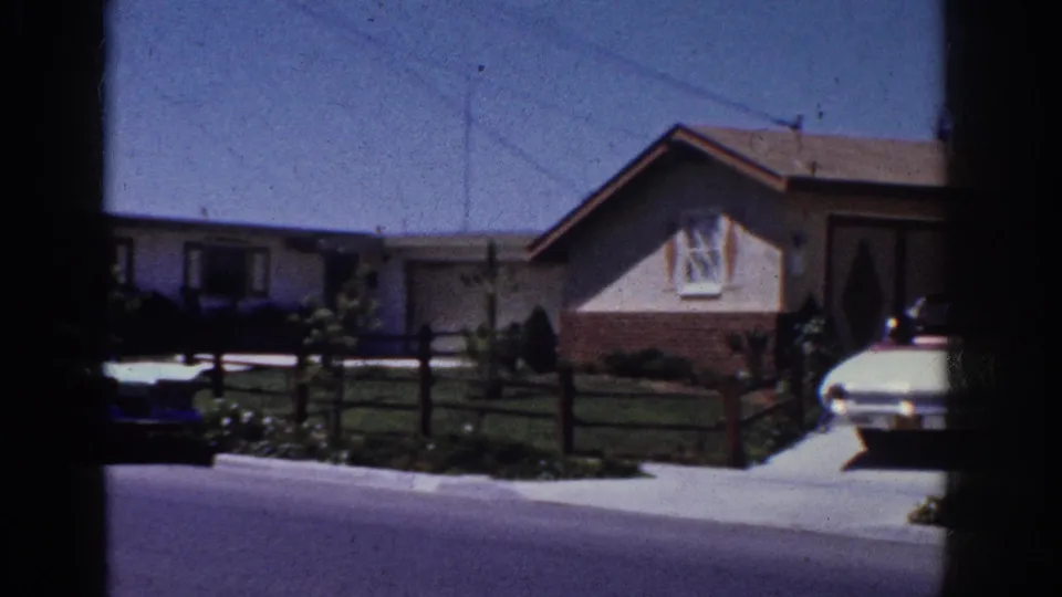 1962 Yorba Linda house frontage with a lawn, low fence, driveway, parked cars, and dark vertical window framing in the foreground.