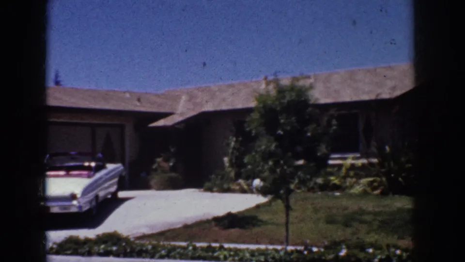 1962 Yorba Linda frame showing the driveway, front lawn, shrubs, small tree, and the white convertible partly cropped at left.