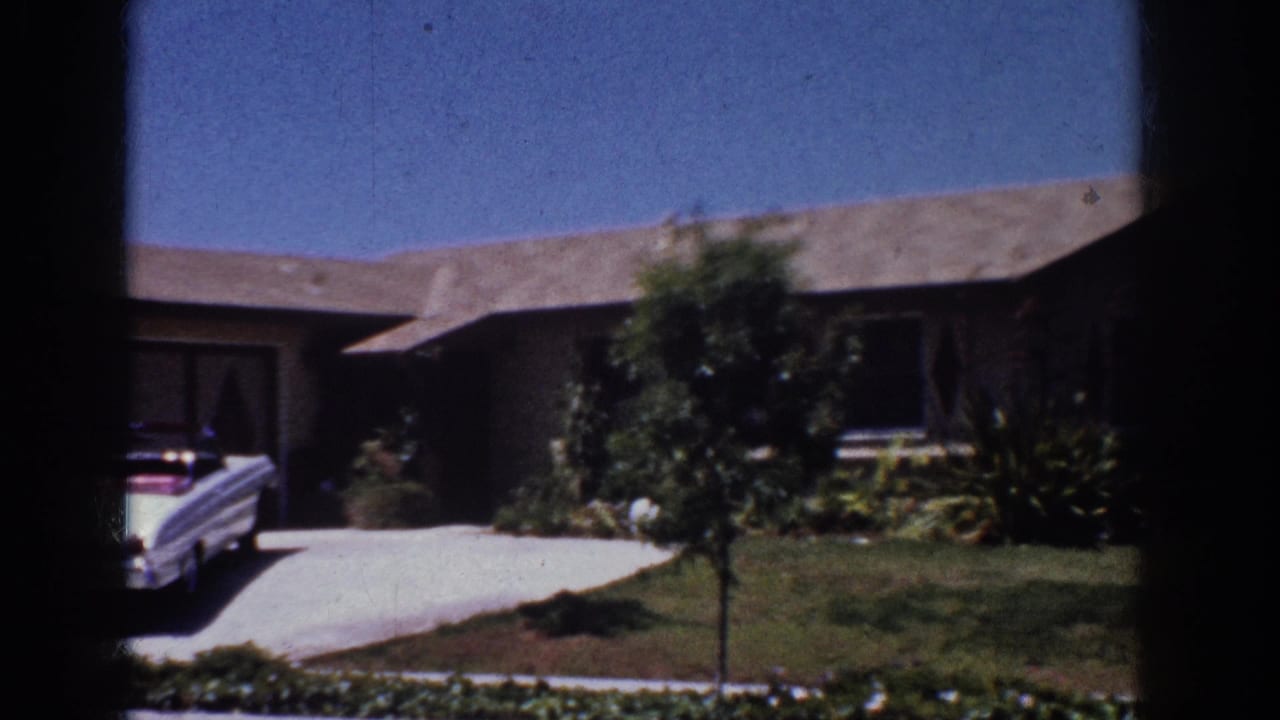 1962 Yorba Linda home-movie frame showing a single-story ranch house, driveway, white car, lawn, shrubs, and blue sky.