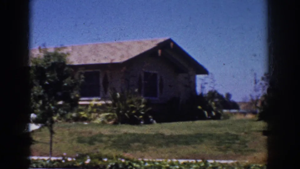 1962 Yorba Linda home-movie frame showing the right side of a ranch house, front lawn, planted shrubs, and a small tree.