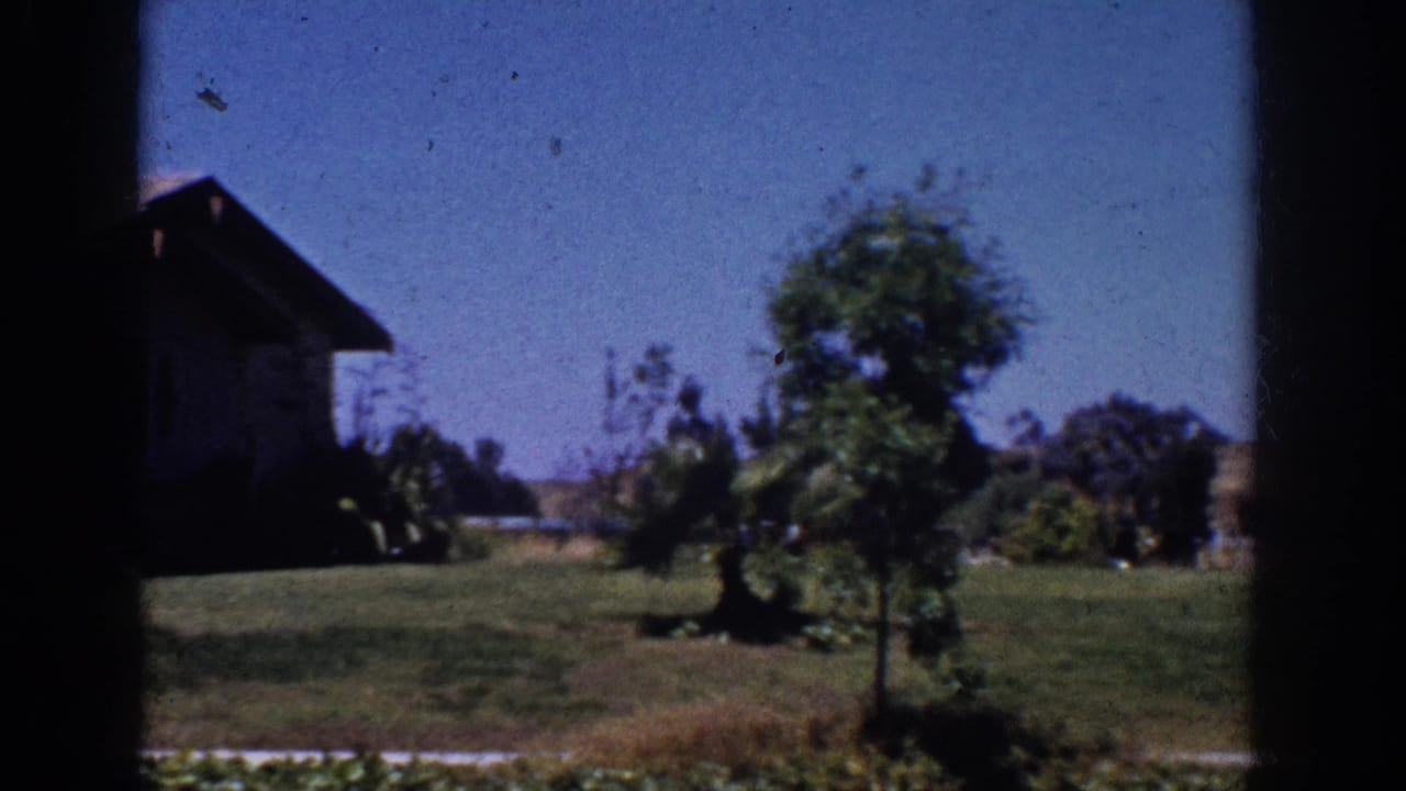 1962 Yorba Linda 8mm frame looking past a dark window edge toward a house, grassy yard, scattered trees, and distant hills.