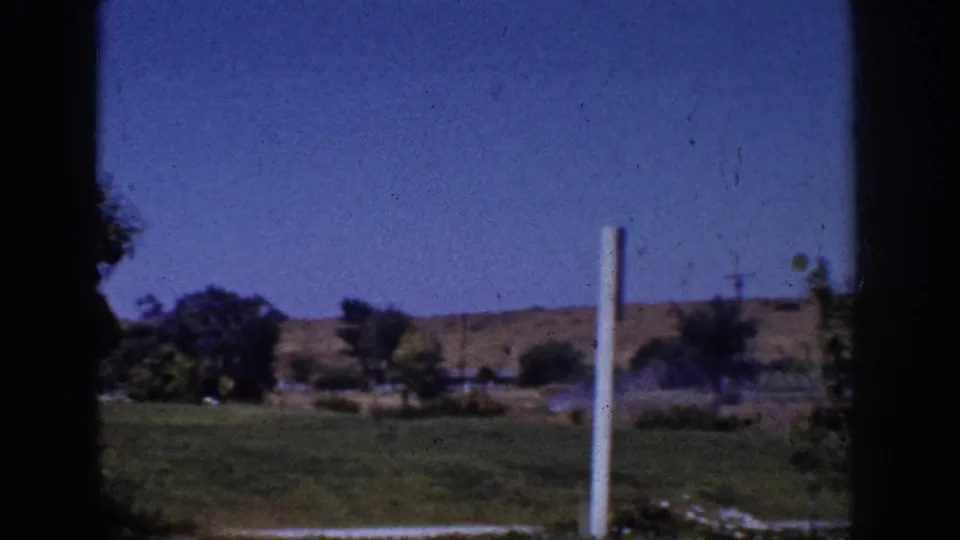 1962 Yorba Linda home-movie frame with open grass, scattered trees, low hills, blue sky, and a pale vertical post in the foreground.