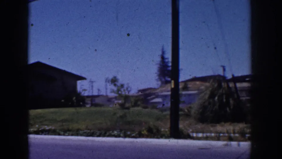 Yorba Linda home-movie frame showing a small dark-roofed building, green lawn, utility poles, trees, and dry hills in 1962.