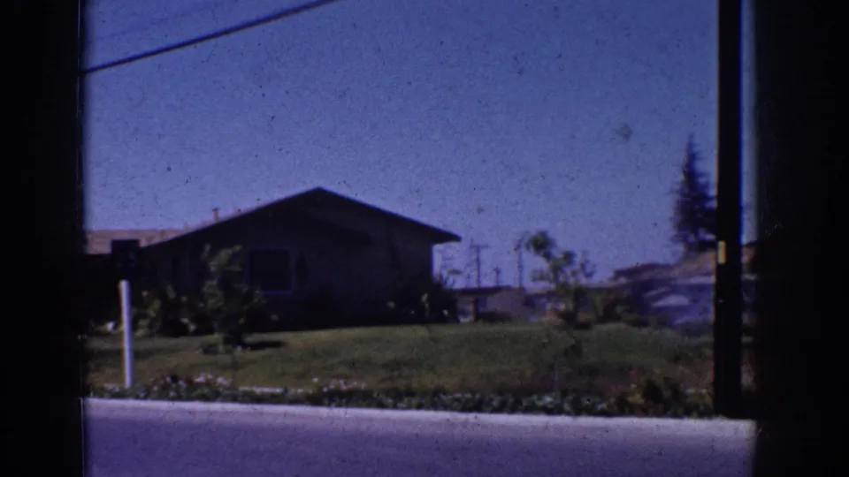 Yorba Linda suburban home exterior with front lawn, roadside post, paved street, shrubs, and a dark vertical edge from the filmed viewpoint.