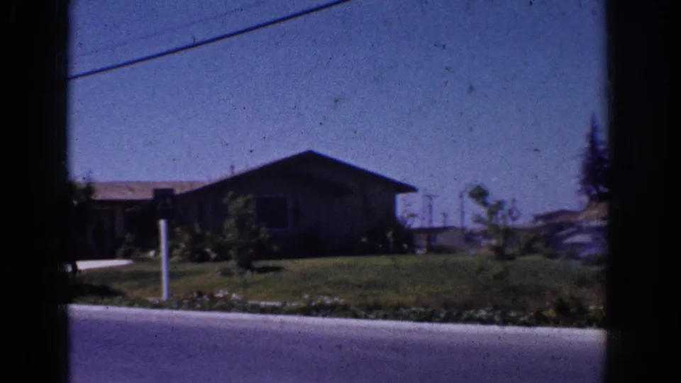 Soft 8mm frame of a 1962 Yorba Linda residential street with a low house, front lawn, curb, white post, and garden plantings.
