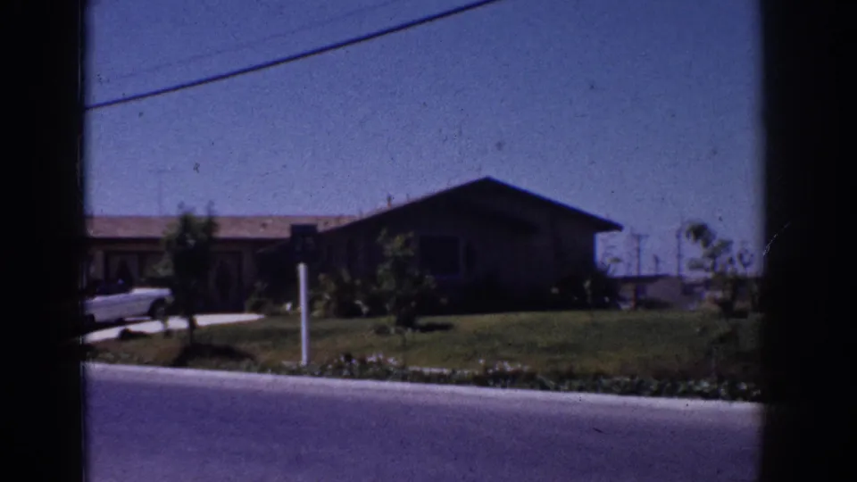 1962 Yorba Linda neighborhood view with a parked white car near a driveway, a single-story house, lawn, shrubs, and street foreground.