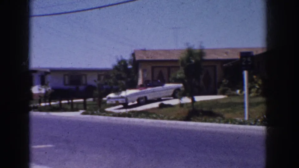 1962 Yorba Linda suburban street frame with a white convertible, driveway, lawn, neighboring houses, and roadside 25 sign.