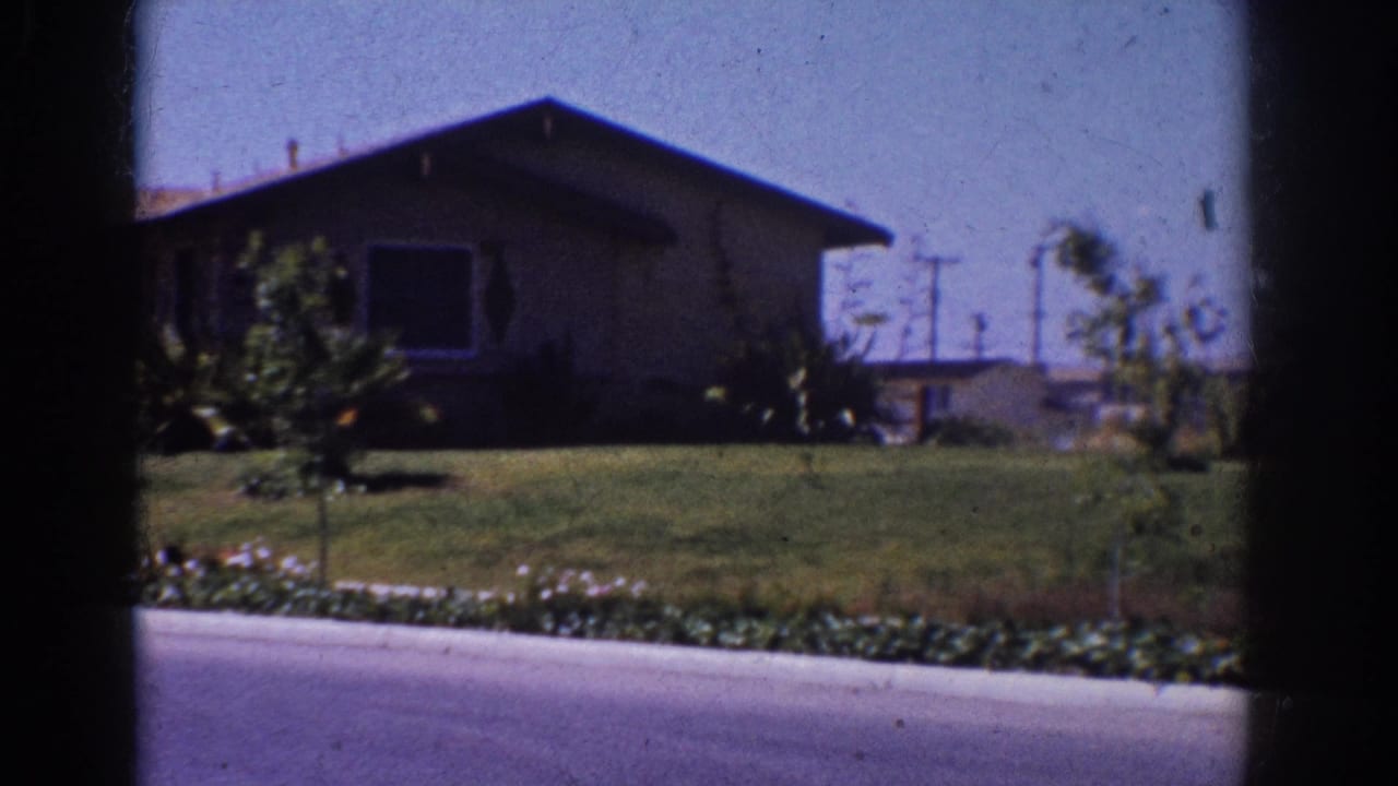 1962 Yorba Linda home-movie frame showing a single-story suburban house, lawn, curb, shrubs, and utility poles from a car-window viewpoint.
