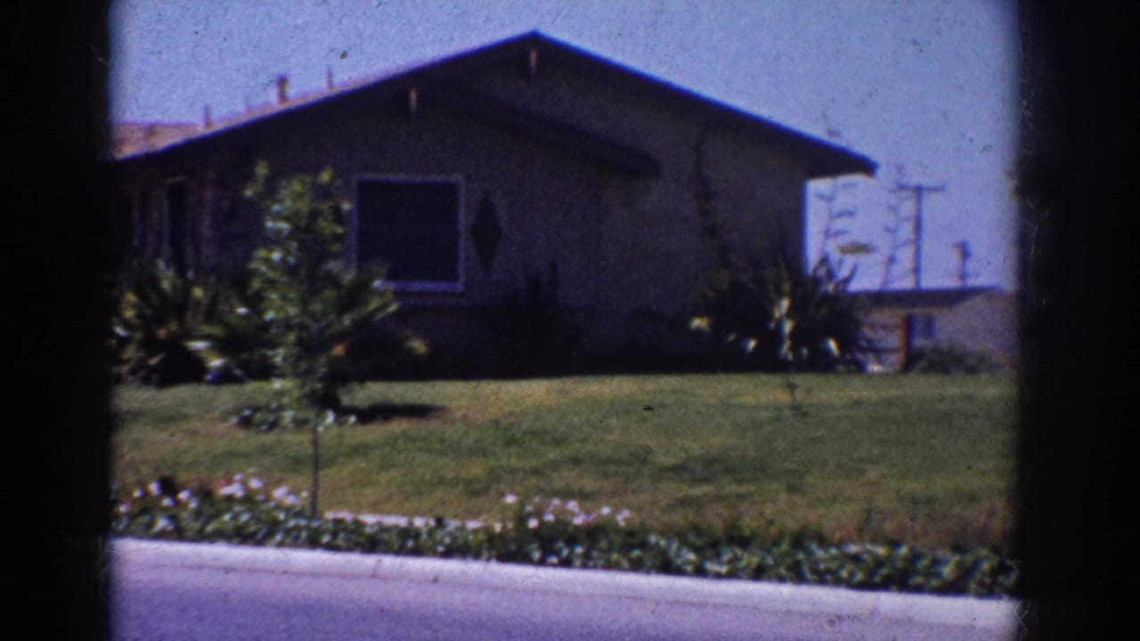 1962 Yorba Linda 8mm frame of a low suburban house, clipped lawn, garden shrubs, and distant utility poles seen from a moving vehicle.