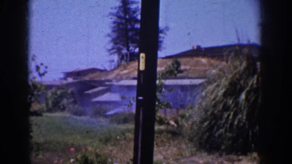 1962 Yorba Linda home-movie frame showing a passing neighborhood view with a dark pole, nearby rooftops, lawn, shrubs, and distant blue sky.