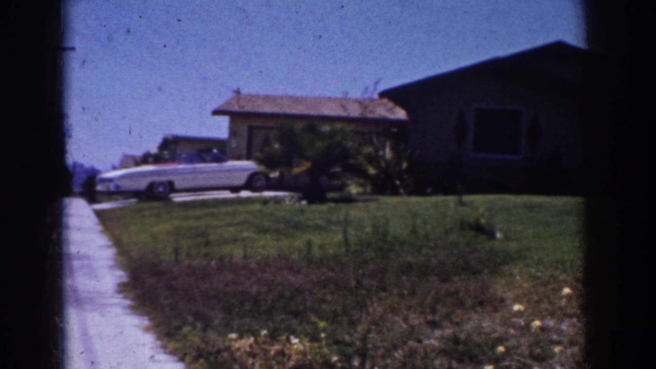1962 Yorba Linda home-movie frame showing a white convertible, sidewalk, lawn, shrubs, and a single-story suburban house.
