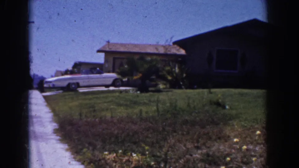 1962 Yorba Linda home-movie frame showing a white convertible, sidewalk, lawn, shrubs, and a single-story suburban house.