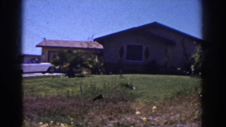 1962 Yorba Linda frame with a single-story home, broad lawn, shrubs, and part of a white convertible at the driveway edge.