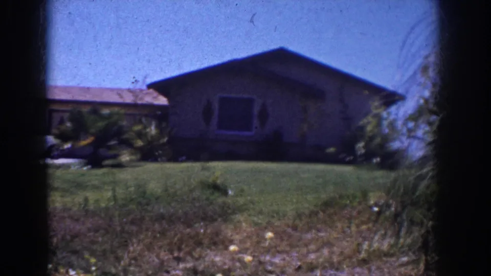 1962 Yorba Linda home-movie frame showing a sloped lawn, house facade, dark front window, shrubs, and soft 8mm gate edges.