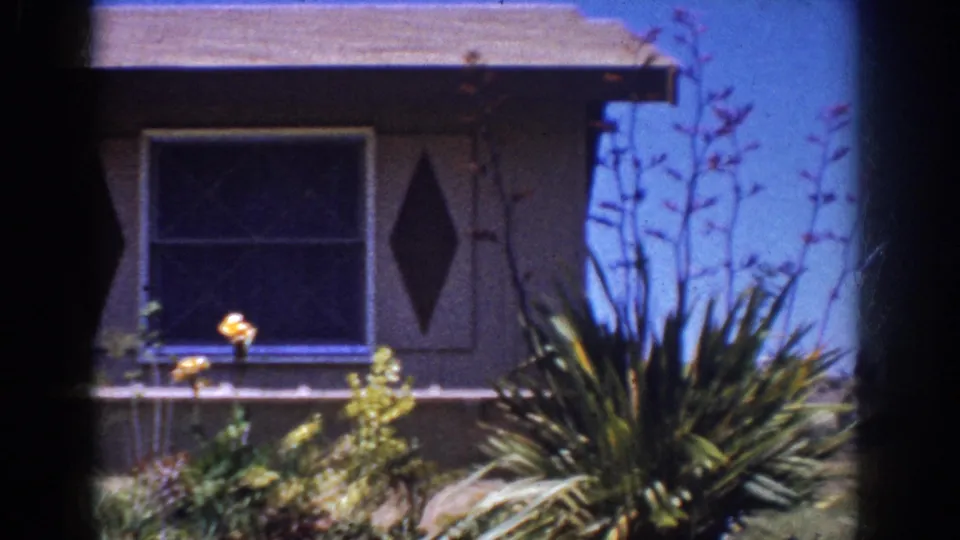 1962 Yorba Linda home-movie frame showing the house corner, large window, diamond shutter, and spiky garden plants.