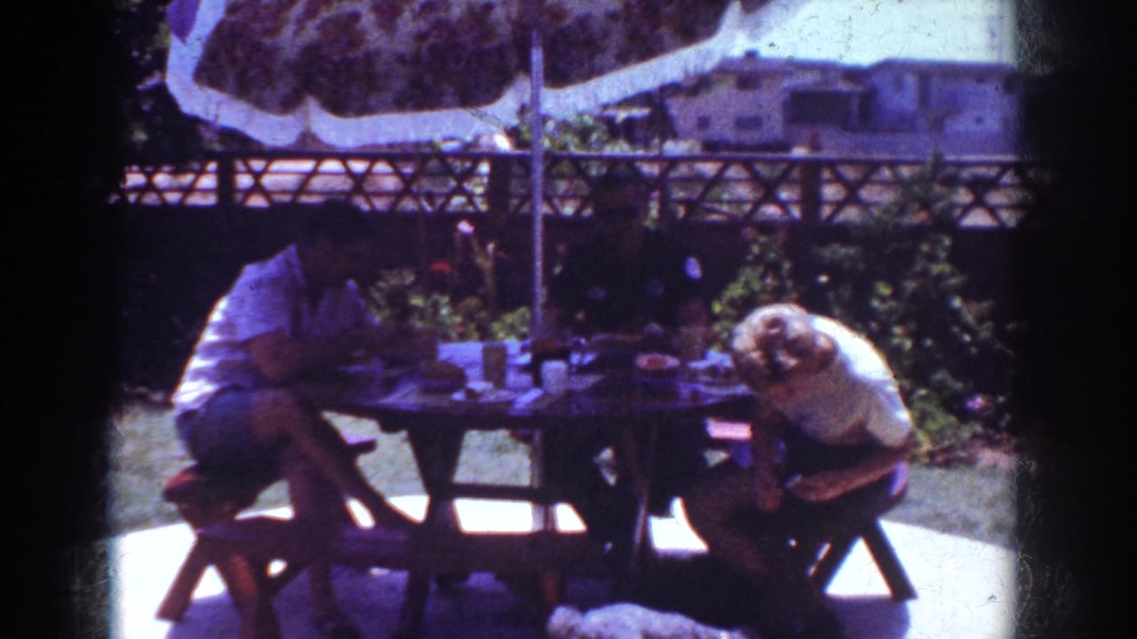 1962 Yorba Linda home-movie frame showing adults at an outdoor picnic table with dishes, umbrella, fence and garden behind them.