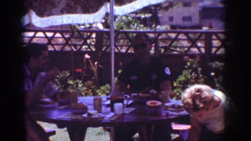 Home-movie frame from Yorba Linda in 1962 showing adults at a backyard table with drinks, plates, umbrella pole and fence.
