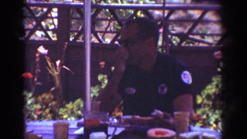 1962 Yorba Linda home-movie frame focused on a seated man in a dark patched shirt at a backyard picnic table.