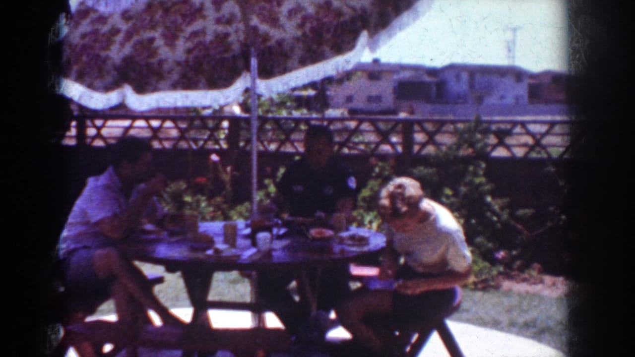 1962 Yorba Linda home-movie frame showing three people around an outdoor picnic table with food, cups, umbrella, fence, and houses beyond.