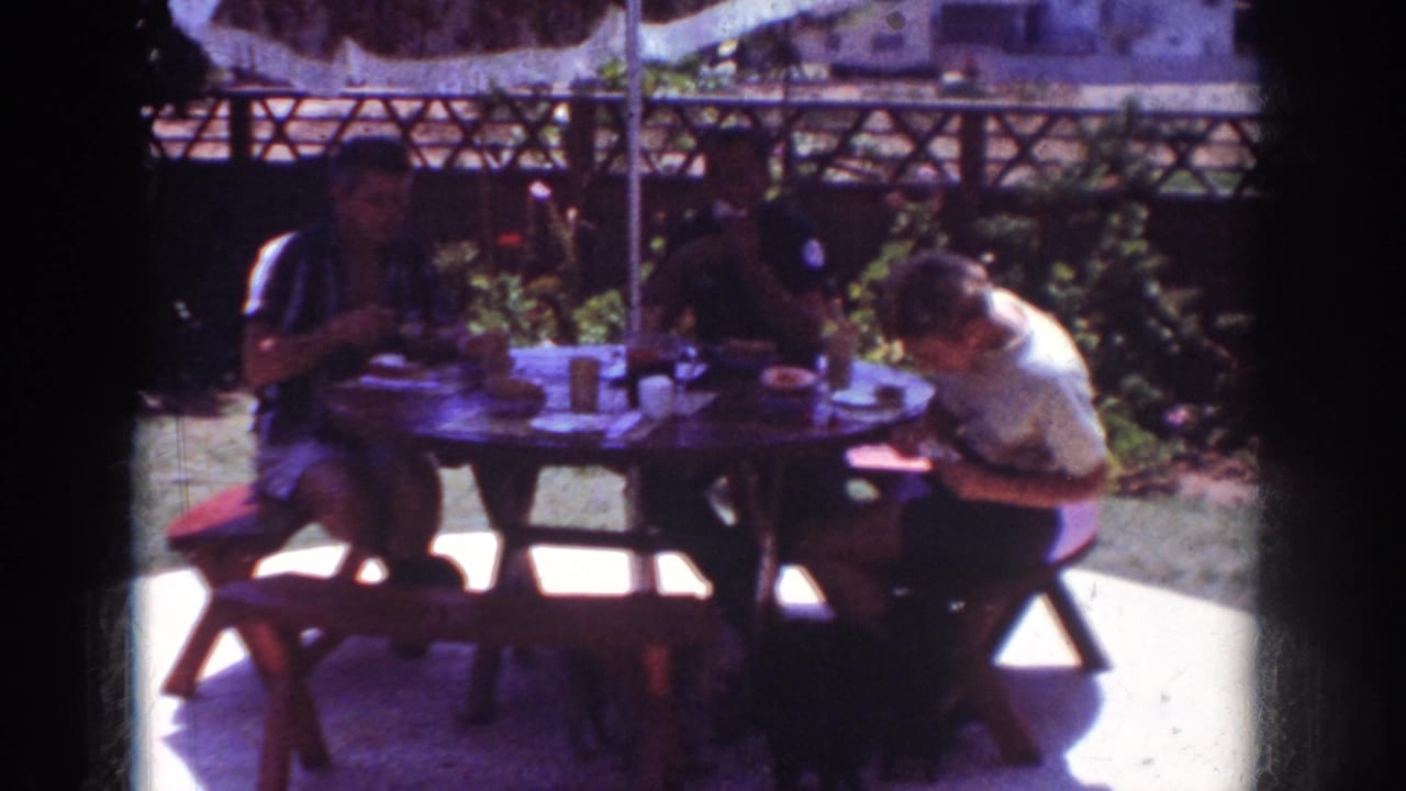 People eat at a shaded backyard table with cups and plates visible.