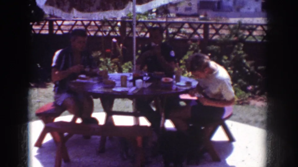 1962 Yorba Linda backyard footage with people seated at a picnic table beneath an umbrella, eating in bright sun and shade.