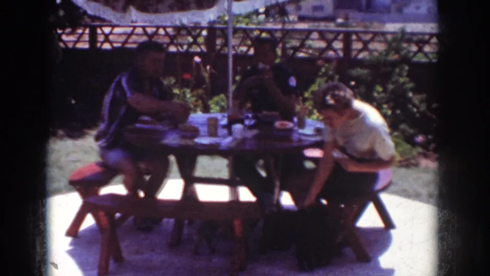 Home-movie frame from Yorba Linda in 1962 showing a close view of people at a backyard picnic table, with garden plants and fencing behind.