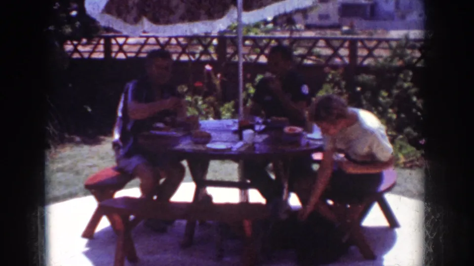 1962 Yorba Linda backyard picnic frame with people eating under a patterned umbrella, surrounded by patio light, plants, fence, and nearby homes.