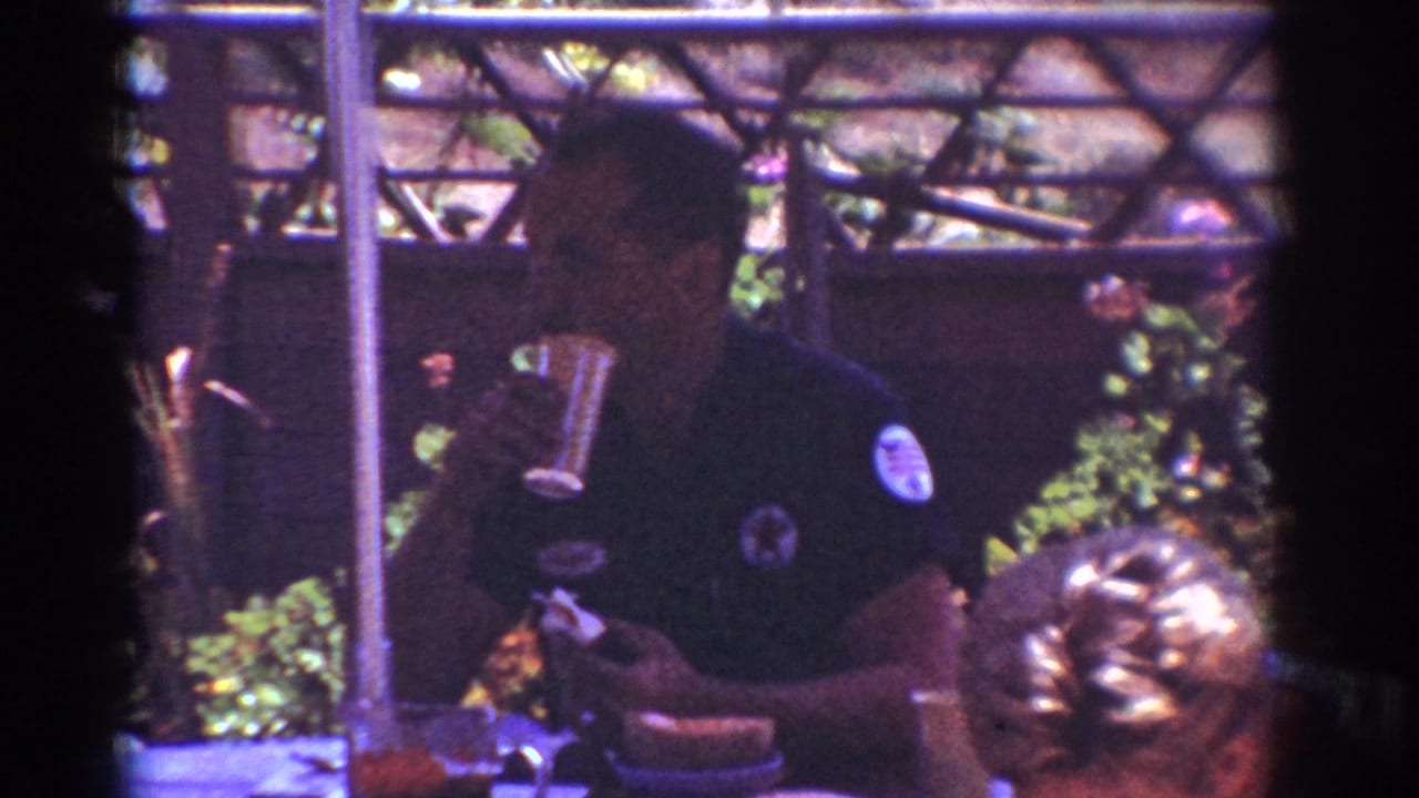 Adult man in a dark shirt holds a glass at a patio table with another person partly visible.
