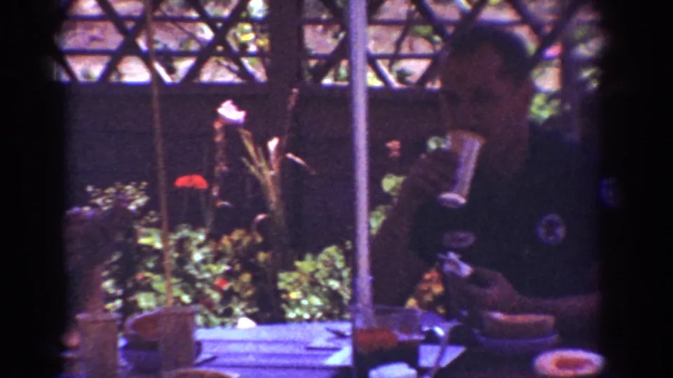 A man in a dark shirt is seen near the patio table, with food, cups, a lattice screen, and plants visible in the 1962 home movie frame.