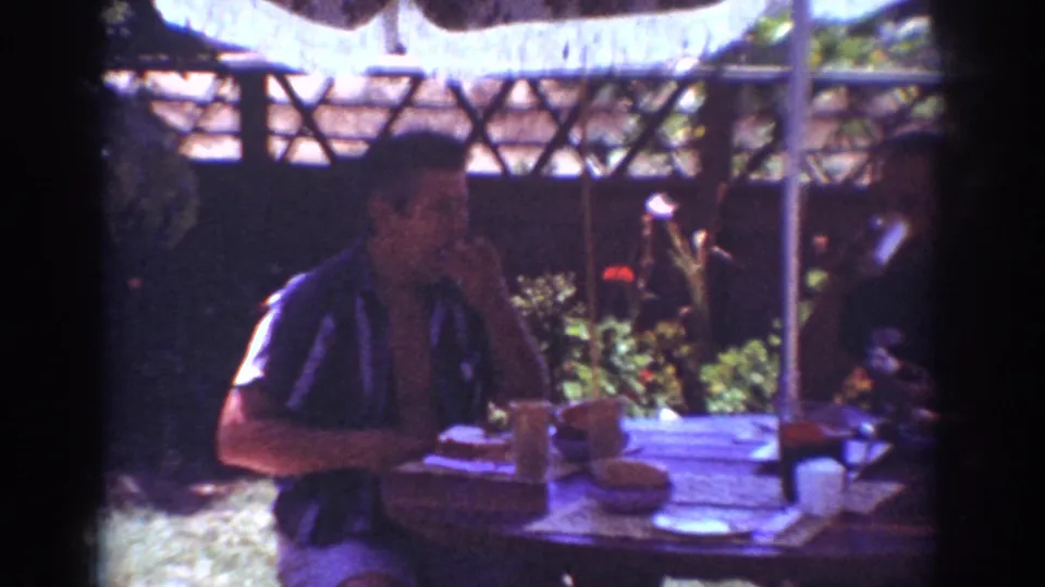 In a wider Yorba Linda patio view, an adult man sits at a round table with food and drinks while another figure is partly visible nearby.