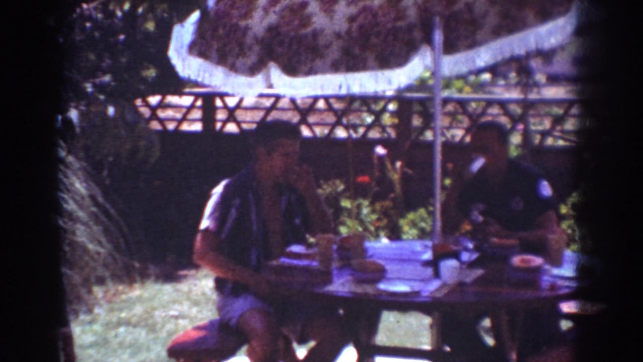 1962 Yorba Linda home-movie frame showing two adult men at a backyard picnic table beneath a fringed umbrella.