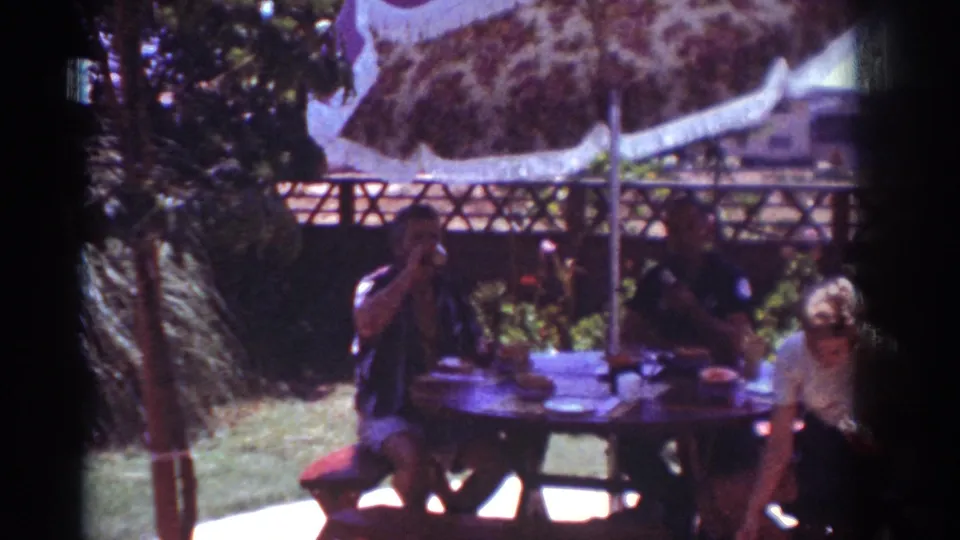 1962 Yorba Linda home-movie frame of adults at a shaded backyard picnic table, with one man raising a drink.