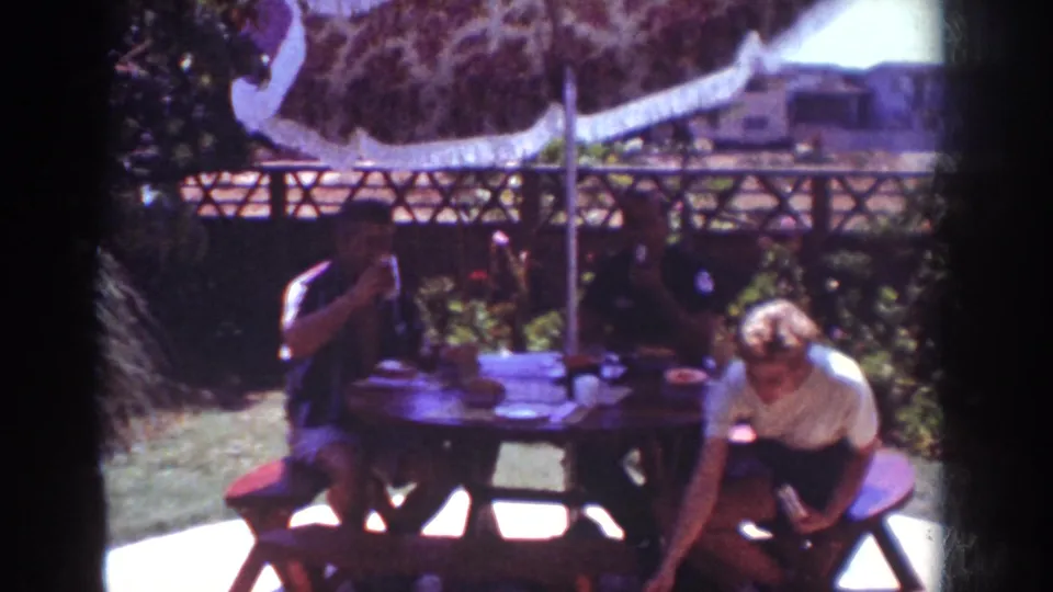 1962 Yorba Linda backyard picnic frame with adults at the table and one person bending beside the bench under the umbrella.