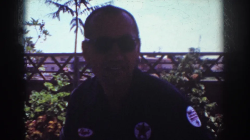 1962 Yorba Linda frame showing a man in sunglasses and a dark shirt with patches facing the camera on a porch with plants and a wooden fence behind him.