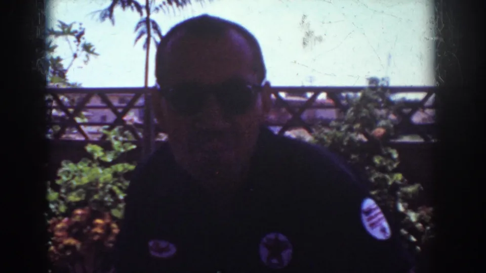 1962 Yorba Linda home-movie frame of a man in sunglasses and a patched dark shirt, backlit against porch plants, a wooden fence, and bright sky.