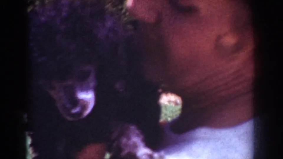A close 8mm frame shows the black puppy next to the woman's face and shoulder, with foliage and lawn visible behind them.