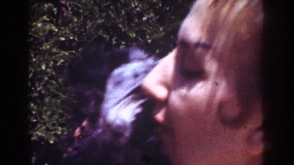 A close 1962 home-movie frame shows the woman kissing or nuzzling the black puppy against a leafy yard background.