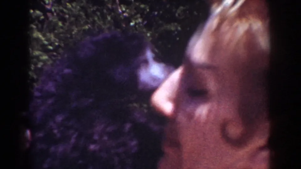 In 1962 Yorba Linda home-movie footage, a woman holds a small dark dog close and kisses its face outdoors.