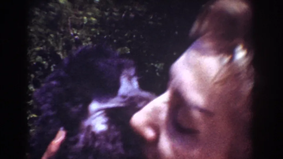 A woman in profile kisses a small dark dog, with foliage behind them and visible softness from the 8mm scan.