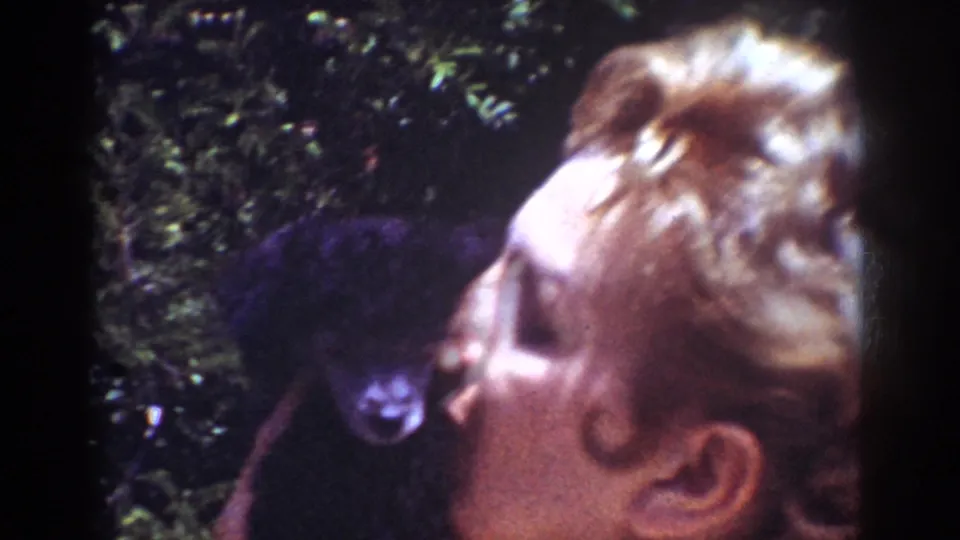 The woman faces a small dark dog at close range in the Yorba Linda home-movie scene, with greenery and soft 8mm texture visible.