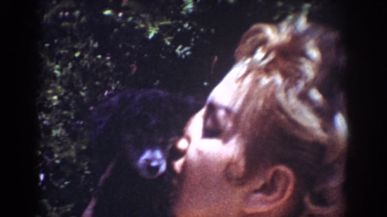 1962 Yorba Linda home-movie frame showing a blonde woman in a white top kissing a small black puppy in a backyard.